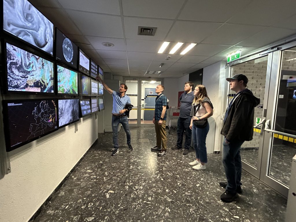 A group of five people standing in a hallway, observing multiple large screens displaying various weather maps and satellite images. One man is pointing to one of the screens, explaining the information being displayed. The group members are casually dressed and appear to be engaged and attentive. The setting is indoors with a tiled floor and overhead fluorescent lighting. The atmosphere suggests an educational tour in a science facility.