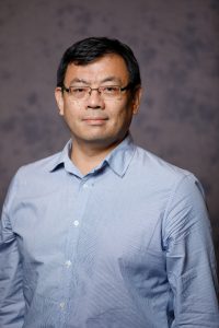 Portrait of a man with short black hair and glasses, wearing a light blue button-up shirt. He has a neutral expression and is standing in front of a gray, slightly textured background.