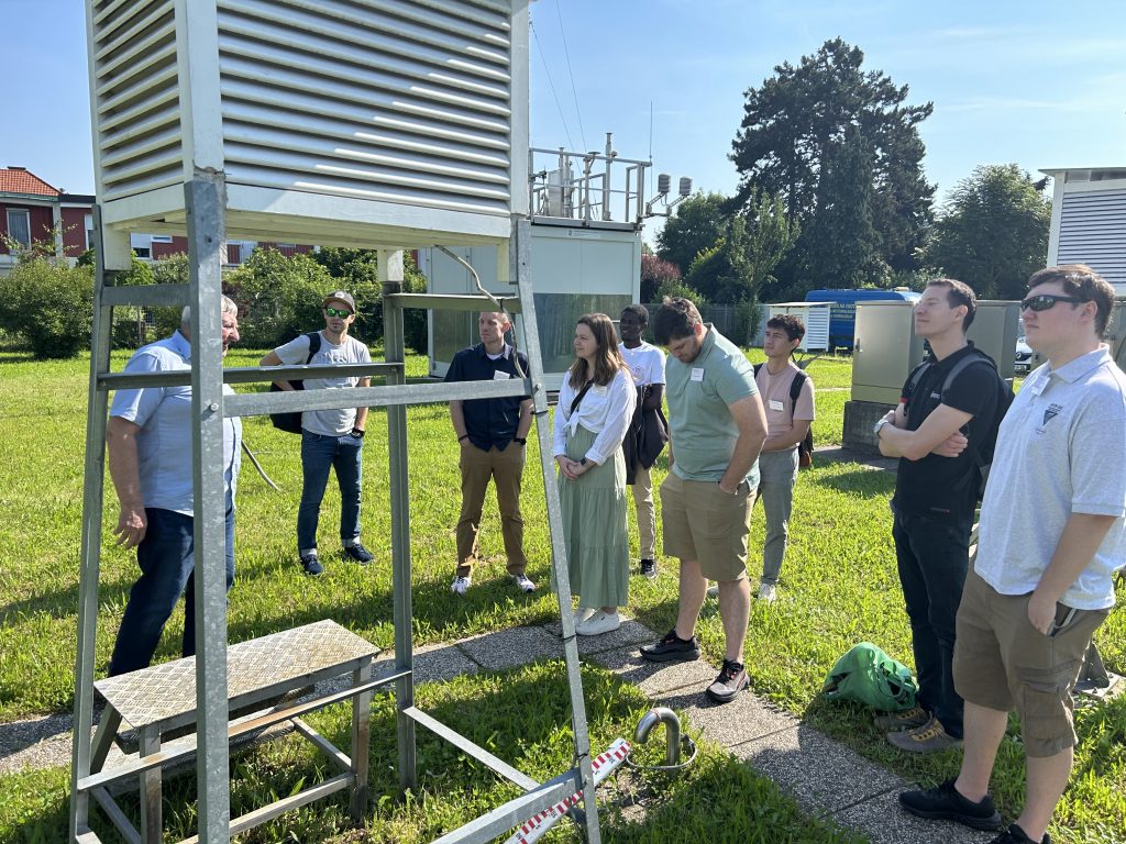 A group of people are standing outside on a grassy area, listening attentively to a man. They are gathered around a metal structure with slatted panels, possibly a weather station or environmental monitoring equipment. The group includes both men and women, dressed casually, with some holding backpacks. In the background, there are various buildings and more equipment, along with trees and a clear blue sky. The setting suggests a field trip or educational tour related to environmental science or meteorology.