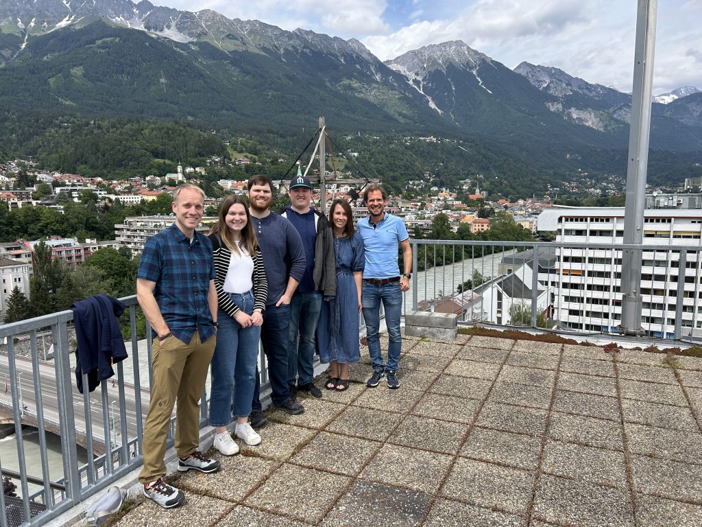 A group of six people is standing on a rooftop with a scenic view of a town nestled in a mountainous area. The individuals are smiling and casually dressed, with a mix of men and women. The town below features various buildings surrounded by lush greenery. In the background, there are towering mountains with patches of snow and forests. The weather appears to be cloudy but bright. The setting suggests a moment of leisure during a work-related trip in a picturesque location.