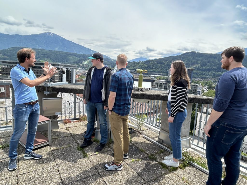 A group of five people is standing on a rooftop, engaged in a discussion. One man, gesturing with his hands, appears to be explaining something to the others, who are listening attentively. The group is casually dressed, and the setting includes various pieces of equipment on the rooftop. In the background, there are buildings, lush green hills, and a mountain with patches of snow under a partly cloudy sky. The setting suggests an educational discussion, related to environmental science, in a picturesque location.