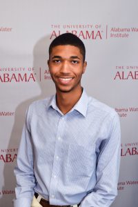 A young man, Kaden Archer, smiling and wearing a light blue checkered button-up shirt. He is standing in front of a backdrop with The University of Alabama and Alabama Water Institute logos. The backdrop is white with red text, and the man is centered in the image, looking directly at the camera.