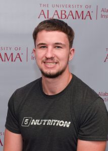 A young man, Peter Feldkamp, smiling and wearing a dark grey t-shirt with the word "NUTRITION" printed on it. He is standing in front of a backdrop with The University of Alabama and Alabama Water Institute logos. The backdrop is white with red text, and the man is centered in the image, looking directly at the camera.