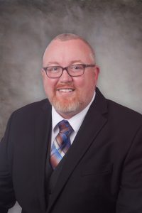 Portrait of a man with short, light-colored hair and a beard, wearing glasses and a dark suit with a white shirt and a striped tie. He is smiling and standing in front of a gray, textured background.