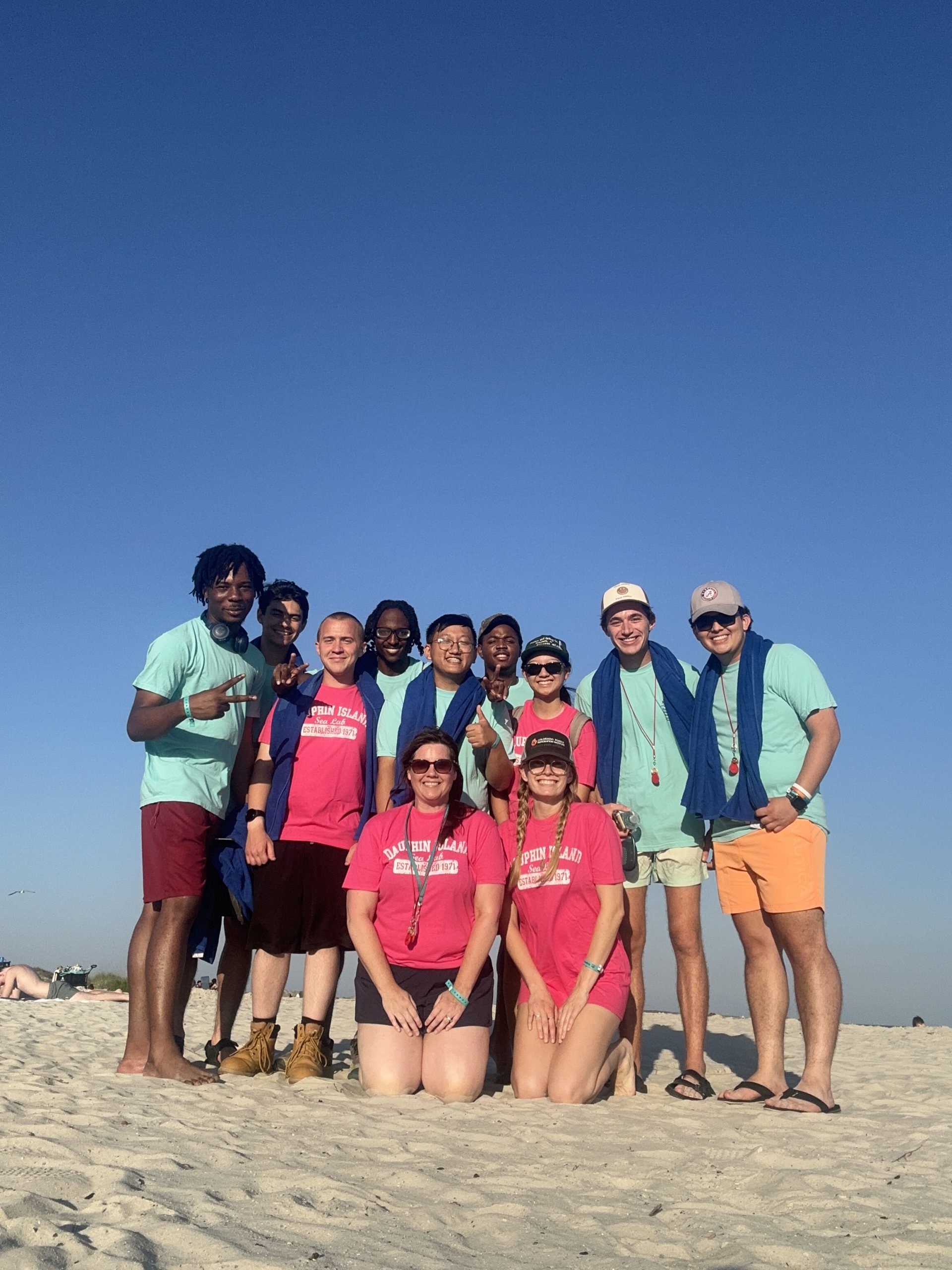 A group of ten people posing on a sandy beach under a clear blue sky. They are participants in the Hydrologic AI Forecasting Program. The group includes both men and women, wearing casual summer clothing in shades of turquoise and pink. Two women are kneeling in the front row, while the rest of the group stands behind them. Everyone is smiling, and some are wearing sunglasses. The background shows a few beachgoers and the expansive sky.