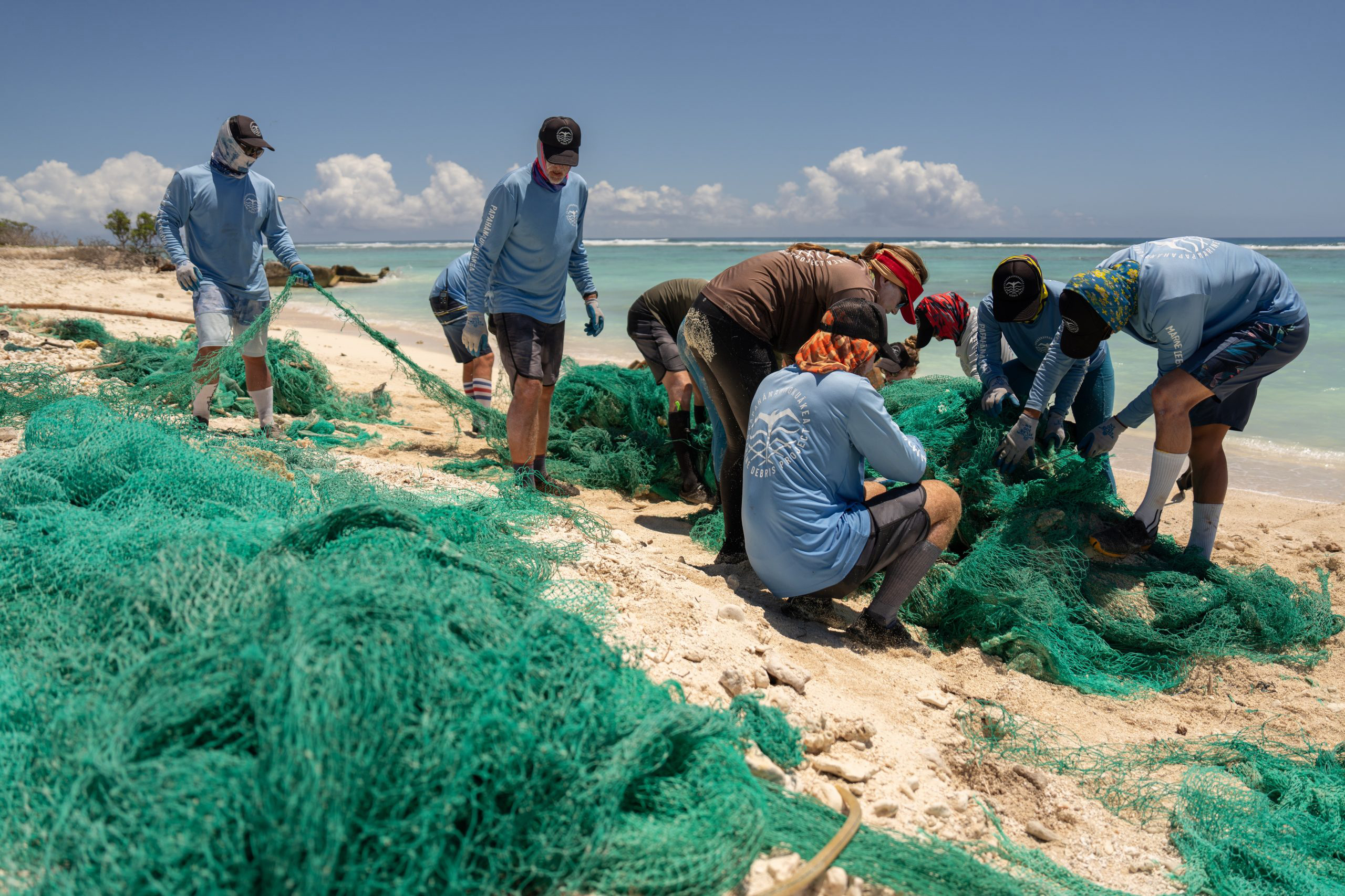 A group of people wearing blue long-sleeve shirts, hats, and gloves are working together to remove large green fishing nets from a sandy beach. The ocean and a clear blue sky are in the background, with some scattered clouds. The scene depicts a coastal cleanup effort.