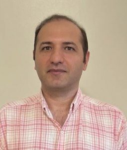 A man with short dark hair, wearing a pink and white checkered button-up shirt, is standing against a plain light-colored background. He is looking directly at the camera with a neutral expression.