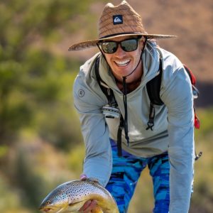 A man wearing a straw hat, sunglasses, and a light gray hoodie is smiling while holding a fish. He has a backpack on and is outdoors, with a blurred natural background indicating a sunny day. The fish appears to be a trout, and the man seems to be enjoying a fishing trip.