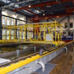 An indoor hydrologic facility featuring a large yellow testing structure suspended over a water channel. The industrial-style building has high ceilings with red pipes and large windows, and people are visible in the background near the testing area.