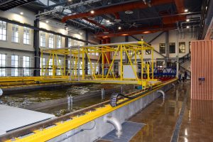 An indoor hydrologic facility featuring a large yellow testing structure suspended over a water channel. The industrial-style building has high ceilings with red pipes and large windows, and people are visible in the background near the testing area.