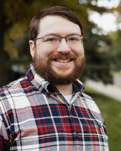 A bearded man with glasses smiles at the camera while wearing a red, white, and blue plaid shirt. He stands outdoors, with a blurred background of green foliage and a hint of autumn colors in the trees.