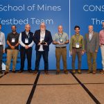 A group of eight men stands on a stage in front of a large blue backdrop with text that reads “Andy Wood, Colorado School of Mines, CIROH Testbed Team” on the left and “Michael Fedoroff, CONSERVE Team, The Wyandotte Nation” on the right. Two individuals in the center are holding awards. Each person is dressed in business casual attire, wearing name badges around their necks. The backdrop also features the CIROH (Cooperative Institute for Research to Operations in Hydrology) logo. The men are positioned in a straight line, facing the camera and smiling.