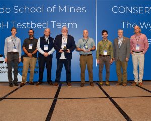 A group of eight men stands on a stage in front of a large blue backdrop with text that reads “Andy Wood, Colorado School of Mines, CIROH Testbed Team” on the left and “Michael Fedoroff, CONSERVE Team, The Wyandotte Nation” on the right. Two individuals in the center are holding awards. Each person is dressed in business casual attire, wearing name badges around their necks. The backdrop also features the CIROH (Cooperative Institute for Research to Operations in Hydrology) logo. The men are positioned in a straight line, facing the camera and smiling.