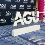 A photograph of the entry hall for the AGU meeting. A freestanding center piece depicting the AGU logo sits in frount of a background with the AGU logo arranged in a checkerboard pattern.