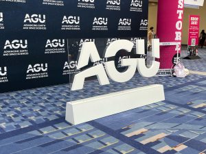 A photograph of the entry hall for the AGU meeting. A freestanding center piece depicting the AGU logo sits in frount of a background with the AGU logo arranged in a checkerboard pattern.