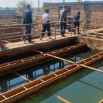 A group of people walking along a concrete walkway at a water treatment facility. The walkway is surrounded by large rectangular basins filled with greenish water. The facility features metal railings and brick buildings in the background under a clear, sunny sky.