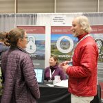 A man in a red jacket and a woman in a purple quilted jacket converse at an exhibition booth for the Alabama Water Institute and CIROH. The booth features banners with logos and images related to water research. A table with informational materials is in the background, where another woman in a purple sweater is seated, smiling. Other attendees and booths are visible in the background at the event.