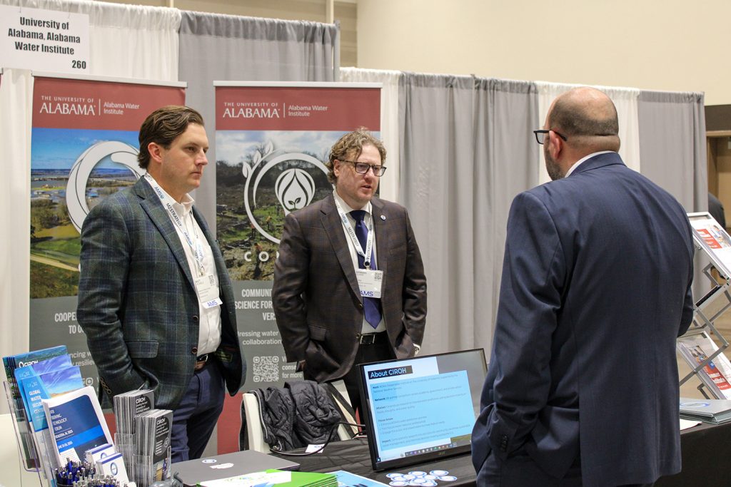 Three men in business attire engage in a conversation at an exhibition booth for the Alabama Water Institute and CIROH. Two of them, wearing lanyards, stand in front of a backdrop featuring The University of Alabama and CIROH logos, while the third man, also in a suit, faces them. A table in the foreground displays brochures, stickers, pens, and a laptop screen showing information about CIROH. A literature stand with informational materials is visible to the right.