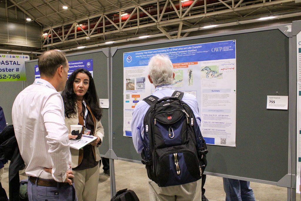 A woman in a beige blazer discusses her research poster with two attendees at a scientific conference. The poster, displayed on a gray board, focuses on water level forecasting for the Great Lakes and Lake Champlain, featuring NOAA and Office of Water Prediction logos. One attendee, a man in a white shirt, listens while holding a cup of coffee, and the other, wearing a blue shirt and a black backpack, faces the poster. The background includes other research posters and attendees in a large convention hall.