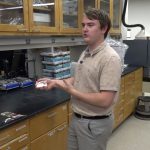 A student wearing a tan shirt stands in a biology lab at a black countertop, holding a slide and explaining his research. Behind him are wooden cabinets, lab equipment, and boxes of supplies.
