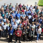 A large group of conference attendees stands outdoors, facing the camera for a group photo on a paved plaza surrounded by grass and trees.
