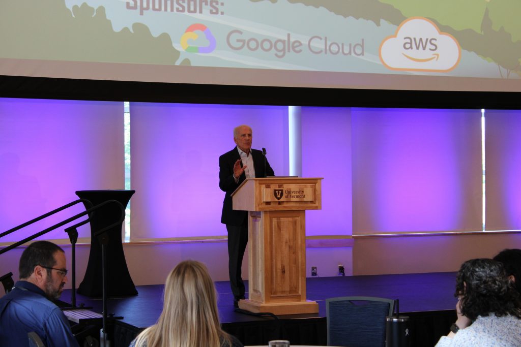 Senator Peter Welch speaks at a wooden podium with the University of Vermont logo, addressing an audience during a keynote session.