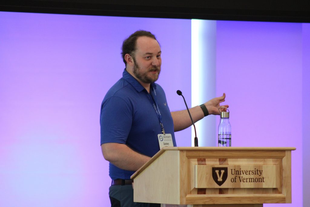 A man speaking at a wooden podium with the University of Vermont logo, addressing an off-camera audience.