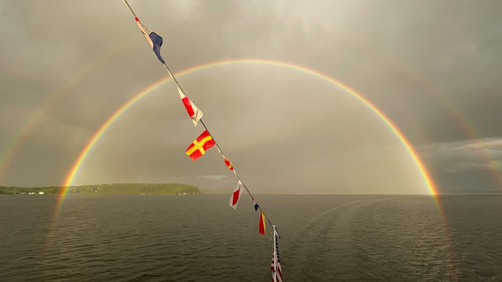 A double rainbow cast above wide-open water, with land and green vegetation visible on the horizon and a series of flags in the foreground.