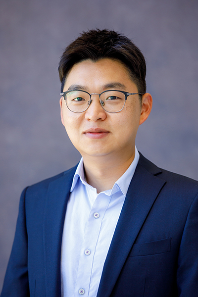 Photograph of Junho Song. Portrait of a man wearing glasses and a dark blue suit with a light blue button-up shirt, standing in front of a neutral gray background.