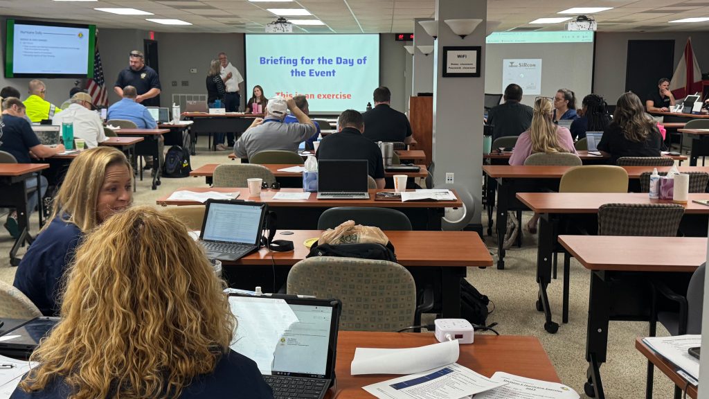 A large classroom with people seated at tables, using laptops and printed materials during a hurricane tabletop exercise. A screen at the front reads “Briefing for the Day of the Event – This is an exercise,” while facilitators and participants engage in discussion.