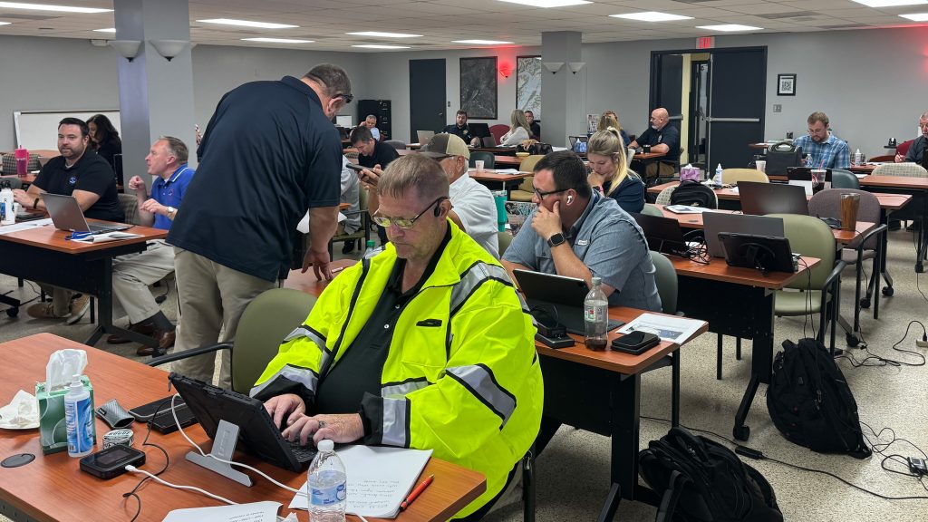 Participants sit at tables with laptops, notebooks, and handouts during the Hurricane Dolly tabletop exercise. One man in a yellow safety jacket types on a tablet while others listen, take notes, or work on computers.