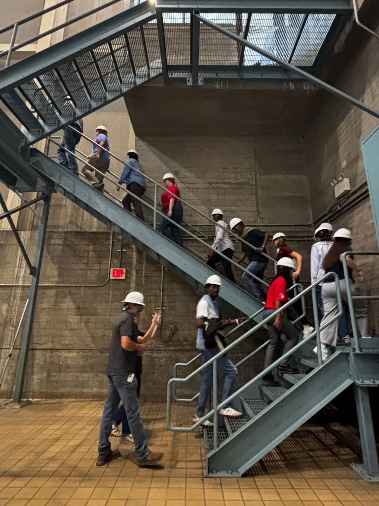 A group of people wearing hard hats walk up a metal staircase inside a concrete dam.