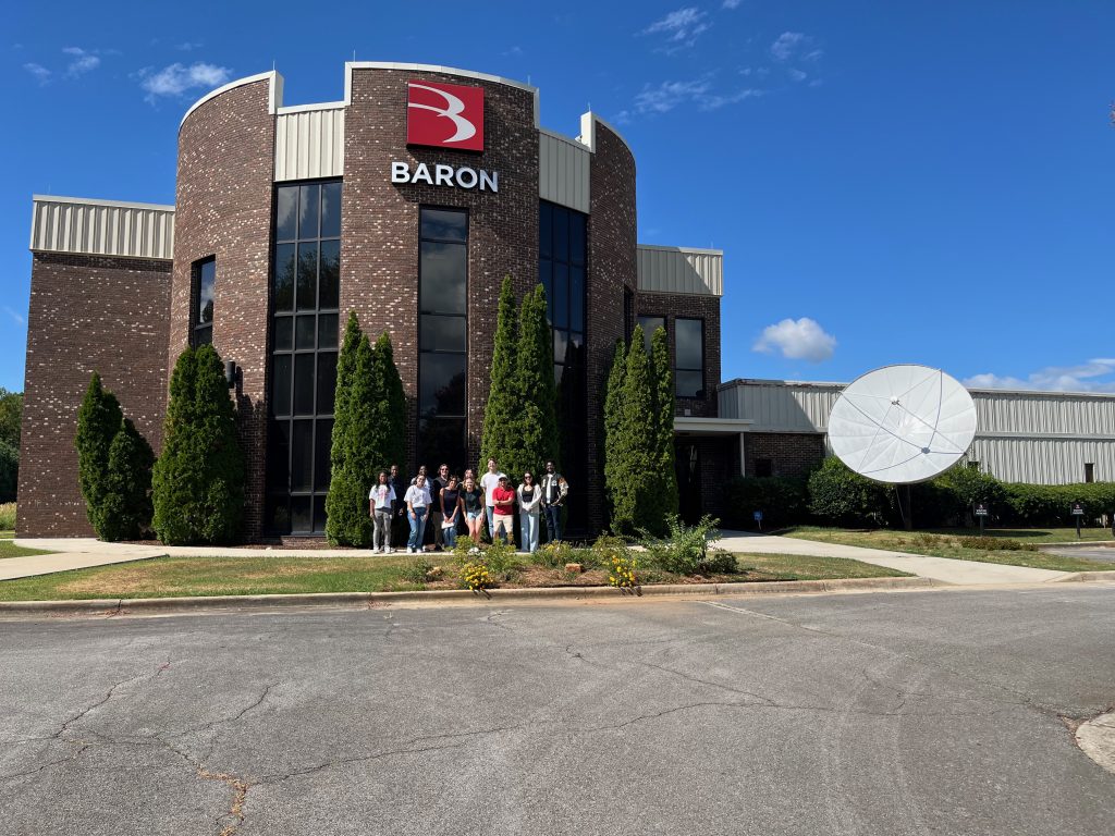 A group of people stand in front of the Baron building with a satellite dish beside it.