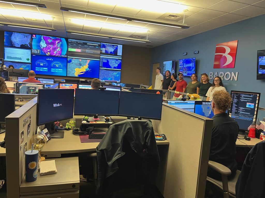 People tour a control room at Baron, with large weather maps displayed on wall monitors.