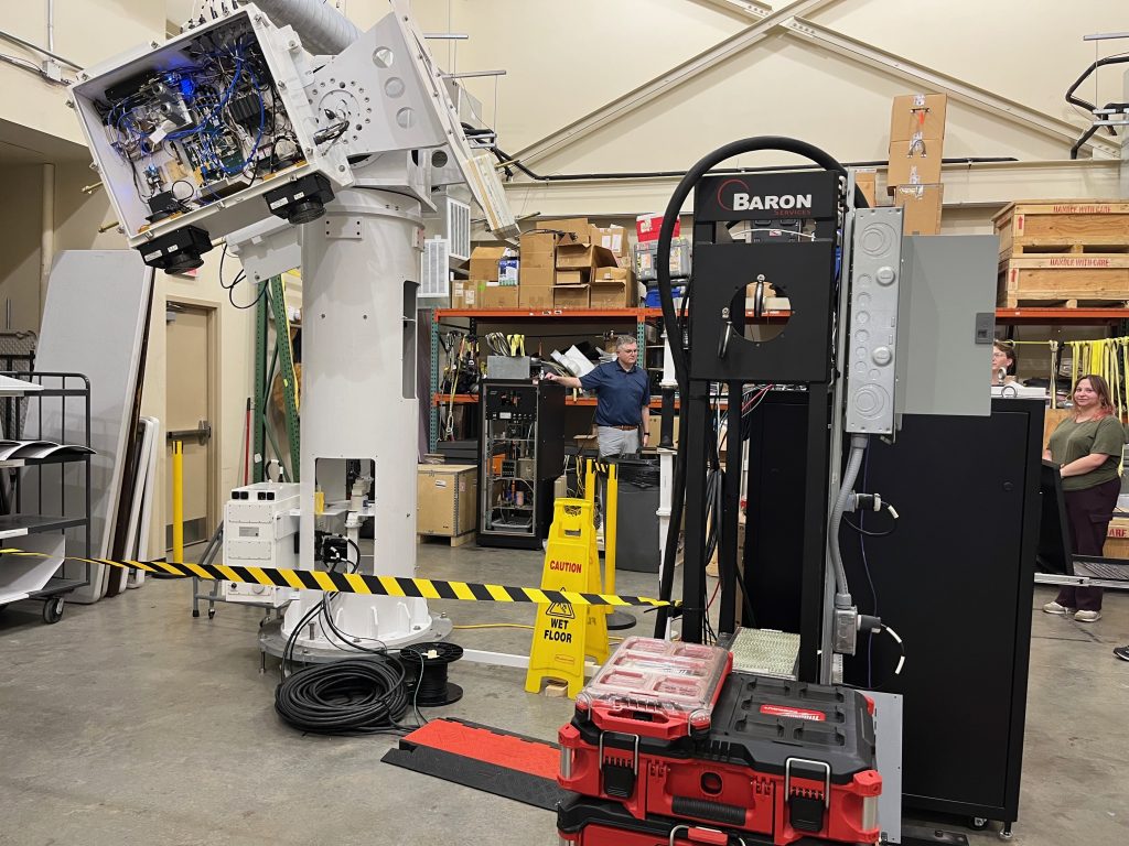 A man speaks to a group beside radar equipment and tools inside a workshop.