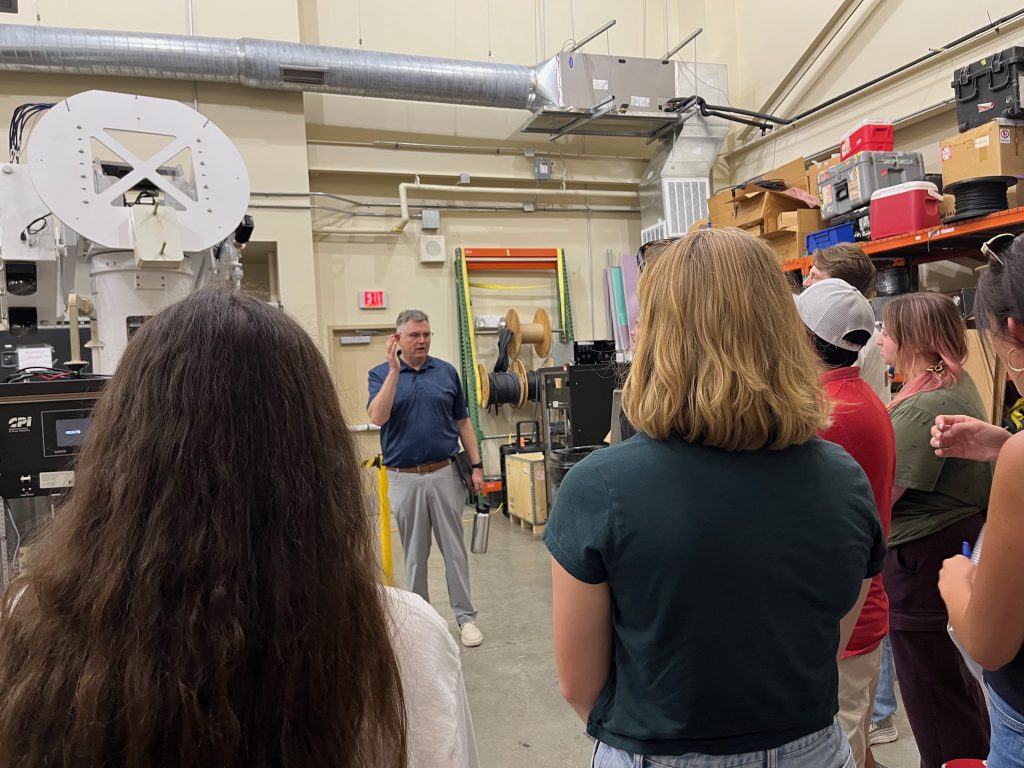 A group listens to a man speaking in a workshop filled with radar equipment and tools.