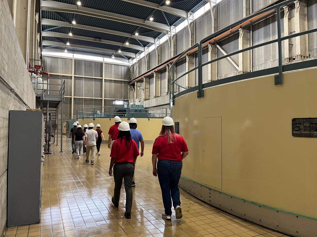 People wearing hard hats walk through a large hall inside a dam facility.