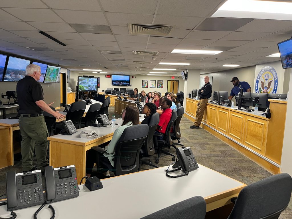 A group listens to a speaker inside an emergency management operations center with large monitors on the wall.
