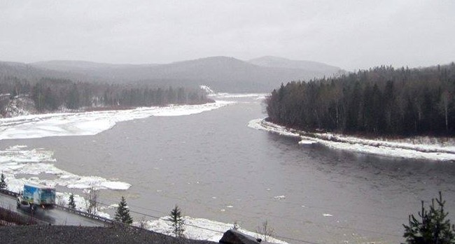 A photo of a river icing over with snow on the riverbanks and a light haze over the midground trees and background mountains.