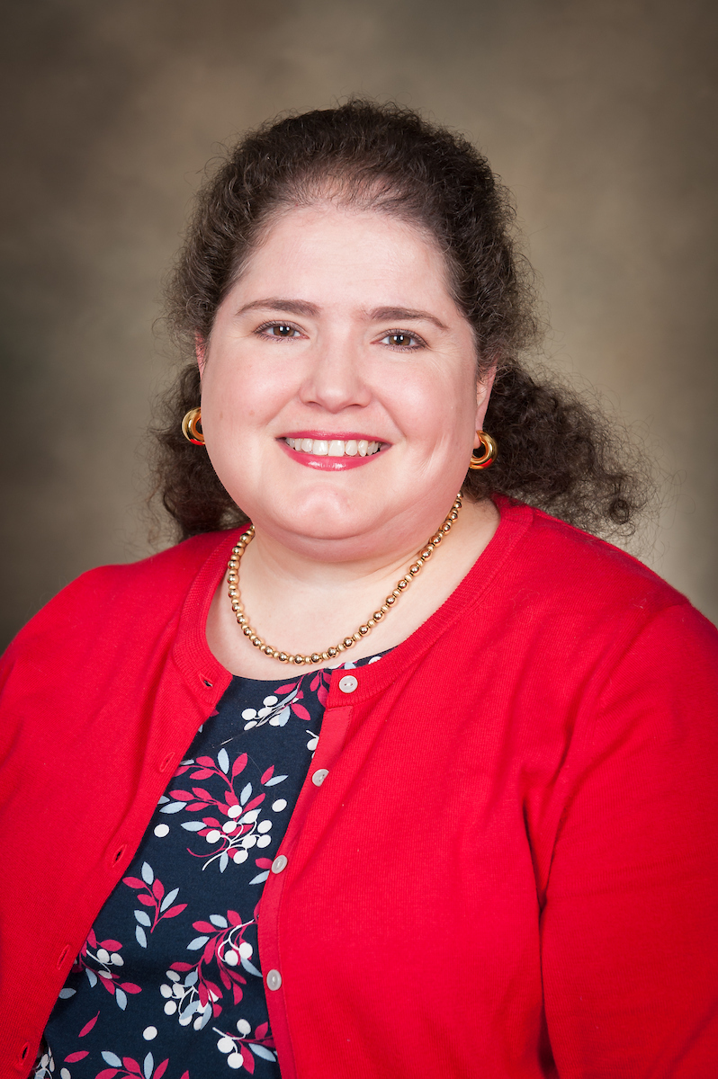 A photo of Dr. Katrina Ramonell in a red sweater and flowered dress in front of a beige background.