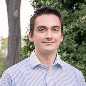 Dr. Adam Hauser stands outdoors near trees, wearing a striped button-down shirt and smiling slightly.
