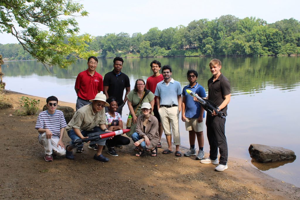 Research students and mentors stand together on a riverbank with hydrology field equipment during a summer research outing.
