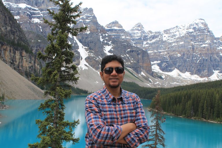 A man poses in front of a turquoise mountain lake surrounded by evergreen trees and snow-covered peaks.