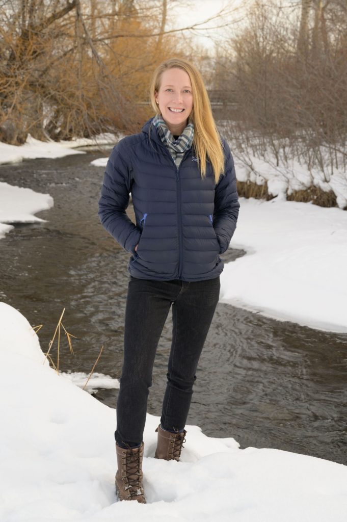 Person standing on snow beside a partially frozen stream, wearing a navy jacket, scarf, black jeans, and brown boots, with bare winter trees in the background.