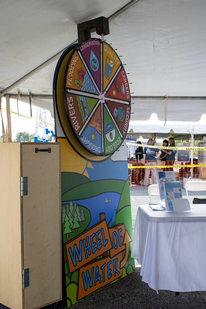 A large, colorful “Wheel of Water” game stands under an event tent, featuring sections on topics like flooding, conservation and ecology. A nearby table holds informational brochures, with attendees visible in the background.