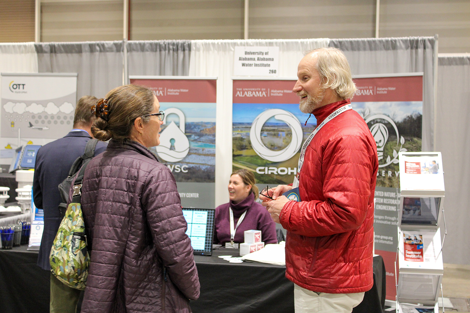 A man in a red jacket and a woman in a purple quilted jacket converse at an exhibition booth for the Alabama Water Institute and CIROH. The booth features banners with logos and images related to water research. A table with informational materials is in the background, where another woman in a purple sweater is seated, smiling. Other attendees and booths are visible in the background at the event.