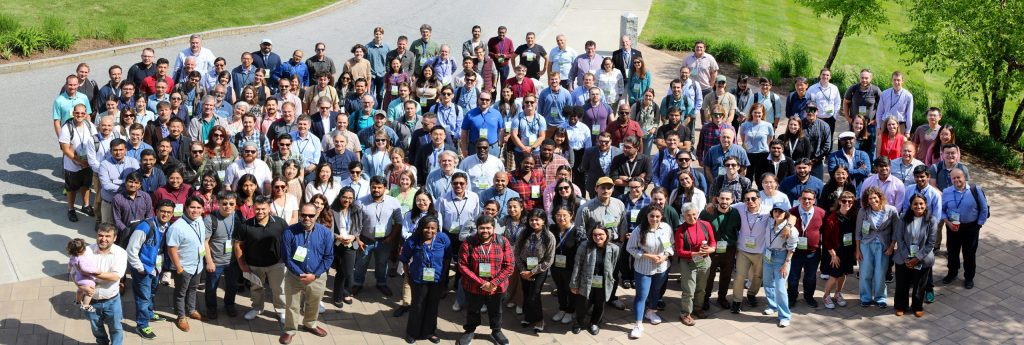 A large group of conference attendees stands outdoors, facing the camera for a group photo on a paved plaza surrounded by grass and trees.