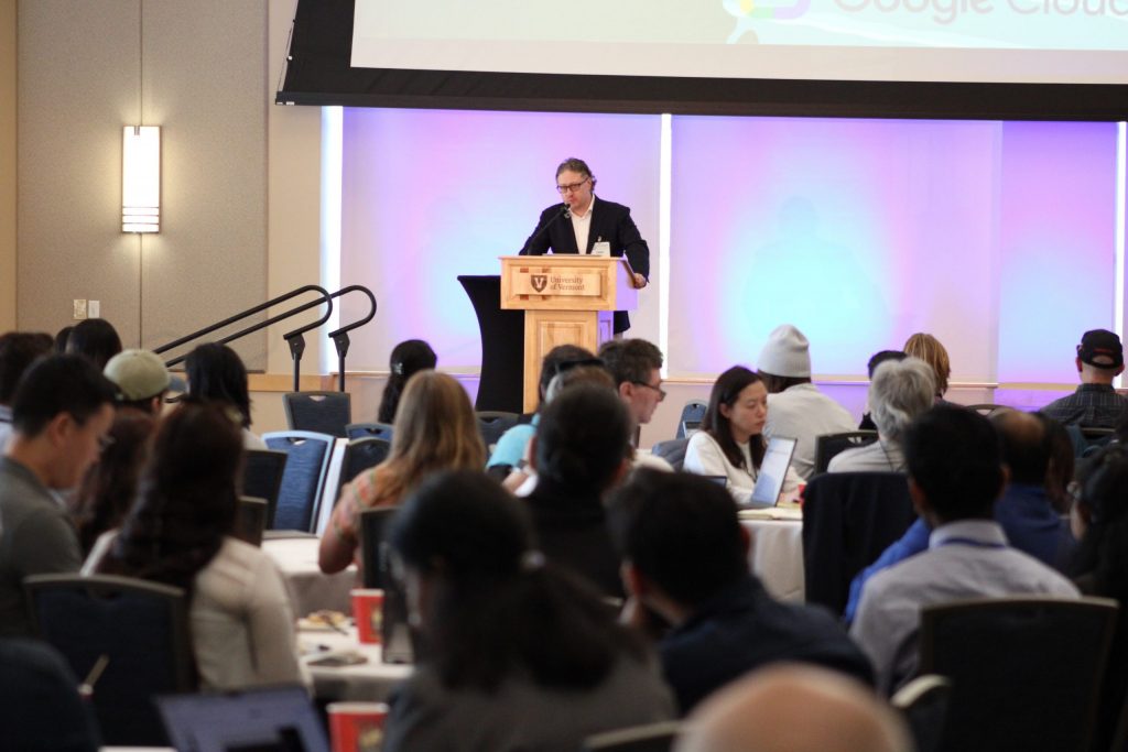 Ed Clark speaks at a wooden podium with the University of Vermont logo, addressing an audience during a keynote session.