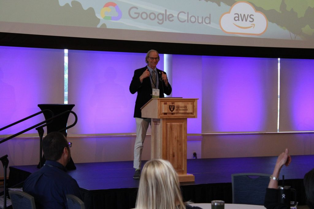 Steve Burian gesturing a double thumbs-up while speaking at a wooden podium with the University of Vermont logo.