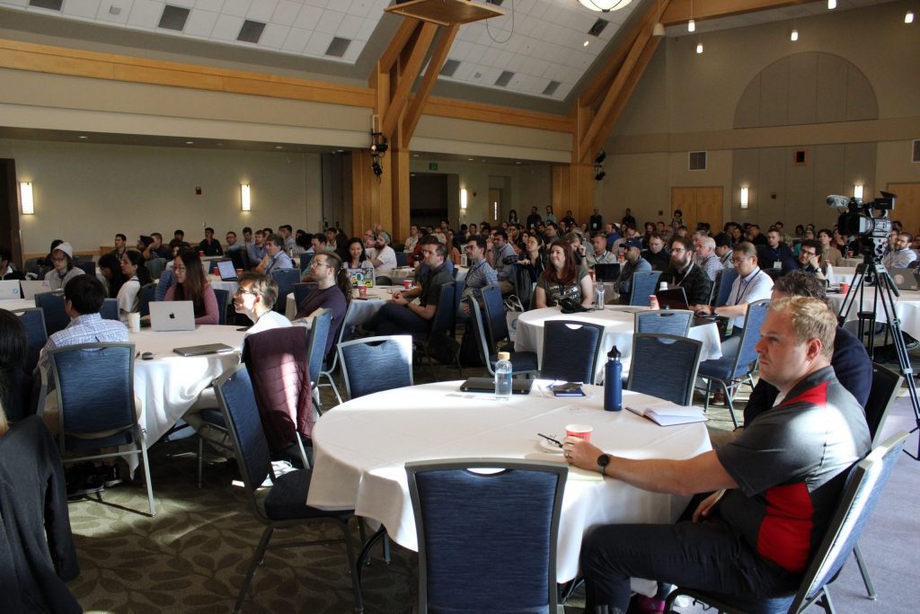 A large conference hall, with numerous conference attendees sitting around several round tables.